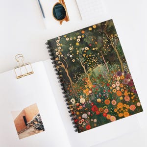 May include: Spiral-bound notebook featuring a colorful floral design with trees and flowers in shades of green, yellow, orange, and red. The notebook is open, revealing blank white pages. A gold-colored paper clip and sunglasses are also visible.
