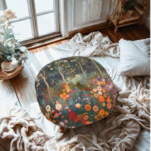 May include: Round floor pillow with a floral design in shades of green, orange, red, and yellow. The pillow has a dark background with a tree-like pattern. The pillow is on a bed with white sheets and a window in the background.