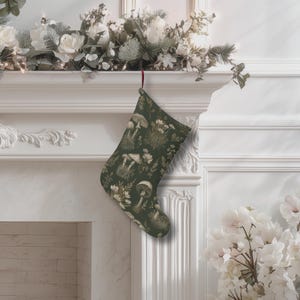 Puede incluir: Un calcetín navideño con un fondo verde oscuro y un estampado de setas y flores. El calcetín cuelga de una repisa blanca decorada con vegetación y flores blancas. El calcetín tiene un lazo rojo.