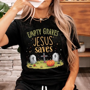 Op de afbeelding: Zwart T-shirt met de tekst "EMPTY GRAVES JESUS SAVES" boven een afbeelding van een kruis, twee grafstenen en een pompoen. Het ontwerp is in groen, oranje en wit.