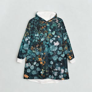 K&ouml;nnte beinhalten: Eine Kapuzen-Decke mit botanischem Druck mit teal-, gr&uuml;nen und orangefarbenen Blumen- und Blattelementen. Der Innenraum und die Manschetten sind wei&szlig;. Die Decke hat eine gro&szlig;e Fronttasche und eine gem&uuml;tliche, &uuml;bergro&szlig;e Passform.