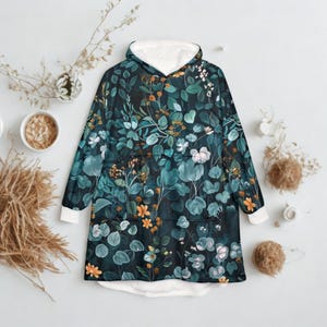 K&ouml;nnte beinhalten: Ein Kapuzen-Decke mit einem botanischen Druck mit teal, gr&uuml;nen und orangefarbenen Bl&auml;ttern auf dunklem Hintergrund. Der Innenraum und die Manschetten sind wei&szlig;. Die Decke hat eine gro&szlig;e Fronttasche und eine gem&uuml;tliche, &uuml;bergro&szlig;e Passform.