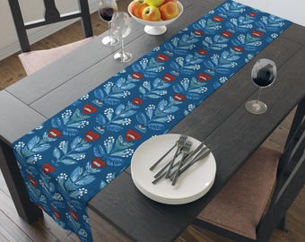 Könnte beinhalten: Blauer Tischläufer mit floralem Muster aus roten Blumen und weißen Beeren. Er liegt auf einem dunklen Holztisch, zusammen mit einer weißen Obstschale, Weingläsern und Tellern mit Gabeln.