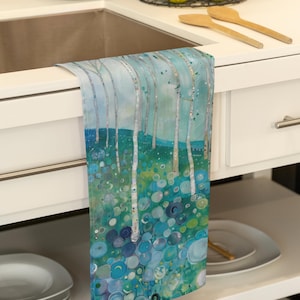 May include: A kitchen towel with a blue and green abstract design featuring a forest scene with trees and circles. The towel is hanging on a white cabinet.