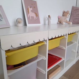 May include: White bookshelf with scalloped edges and yellow storage bins. The bookshelf has two shelves and is decorated with a rainbow picture, a snow globe, and a unicorn picture. The text on the rainbow picture reads "Be Happy".