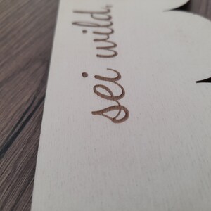 May include: A white wooden sign with a scalloped edge. The sign has the words "sei wild" written in brown cursive.