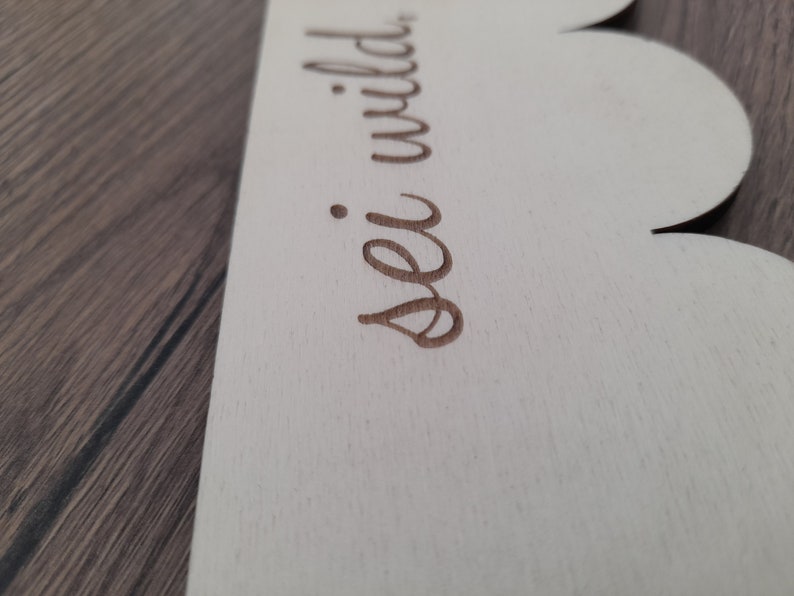 May include: A white wooden sign with a scalloped edge. The sign has the words "sei wild" written in brown cursive.