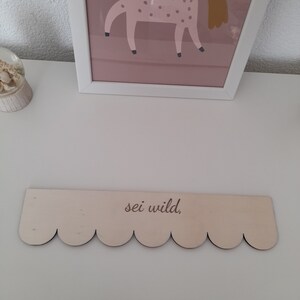 May include: A wooden sign with a scalloped edge and the text "sei wild." The sign is light brown and has a natural wood finish.