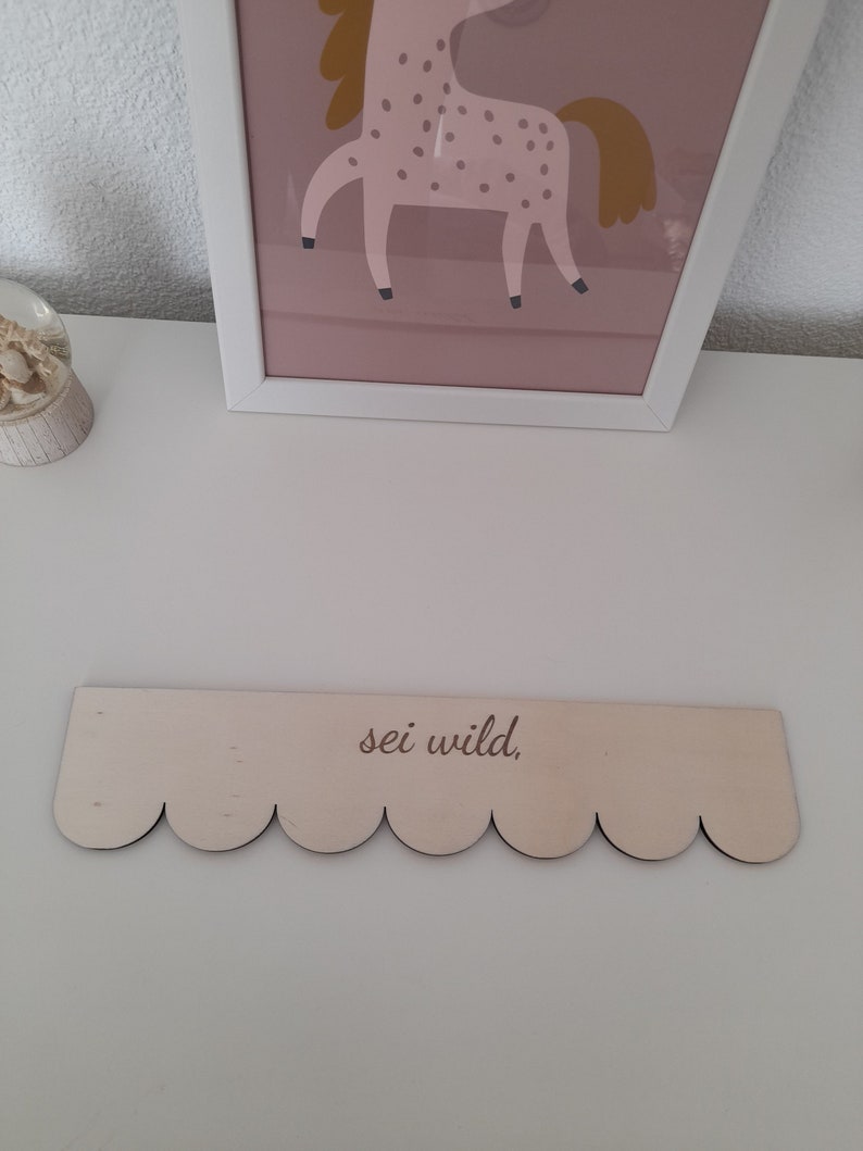 May include: A wooden sign with a scalloped edge and the text "sei wild." The sign is light brown and has a natural wood finish.
