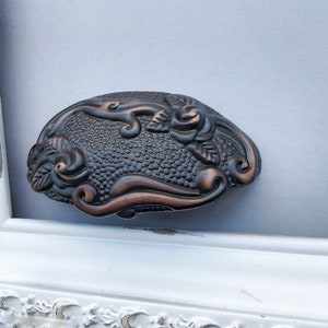 May include: A decorative metal hair clip with a floral design. The clip is a dark bronze color with a textured surface. The floral design features a rose and leaves.