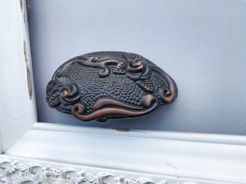 May include: A decorative metal hair clip with a floral design. The clip is a dark bronze color with a textured surface. The floral design features a rose and leaves.