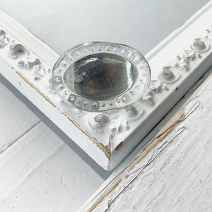 May include: A clear glass oval-shaped knob with a beaded edge, set against a white painted frame with ornate details.