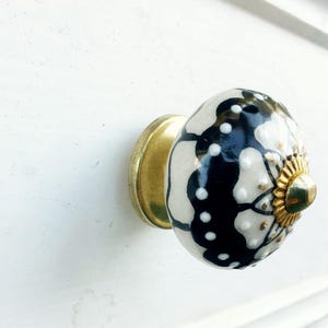 May include: Black and white ceramic knob with a gold accent. The knob has a floral design and is mounted on a white surface.