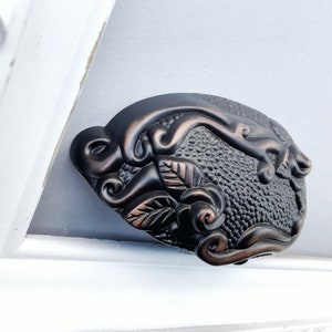 May include: A decorative cabinet pull with a floral design. The pull is made of metal and has a dark bronze finish.