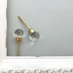 May include: Two clear glass ball cabinet knobs with brass hardware and threaded rods. The knobs are set against a light blue background, with a white distressed picture frame in the corner.