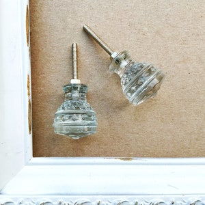 May include: Two clear glass cabinet knobs with decorative detailing. Each knob has a threaded metal rod for installation. The knobs are on a neutral background, suggesting a vintage or antique aesthetic. The knobs are likely for furniture or cabinetry.