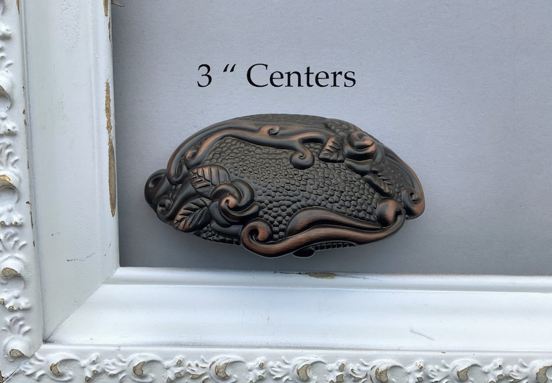 May include: A decorative, ornate, bronze-colored cabinet pull with a floral design. The pull is 3 inches in diameter. The text "3" Centers" is visible above the pull.