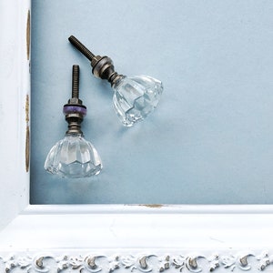 May include: Two clear glass cabinet knobs with a faceted design and brass accents. The knobs are attached to a white painted frame.