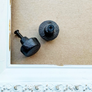 May include: Two black metal cabinet knobs with a textured finish. One knob is round and the other is a more geometric shape.