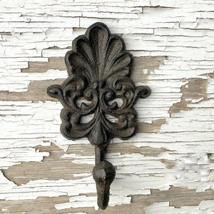 May include: Black cast iron hook with a decorative floral design. The hook is mounted on a white painted wooden surface.
