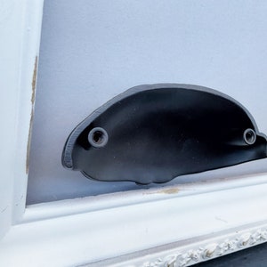 May include: A black metal door handle with two visible screws. The handle is mounted on a white painted frame.