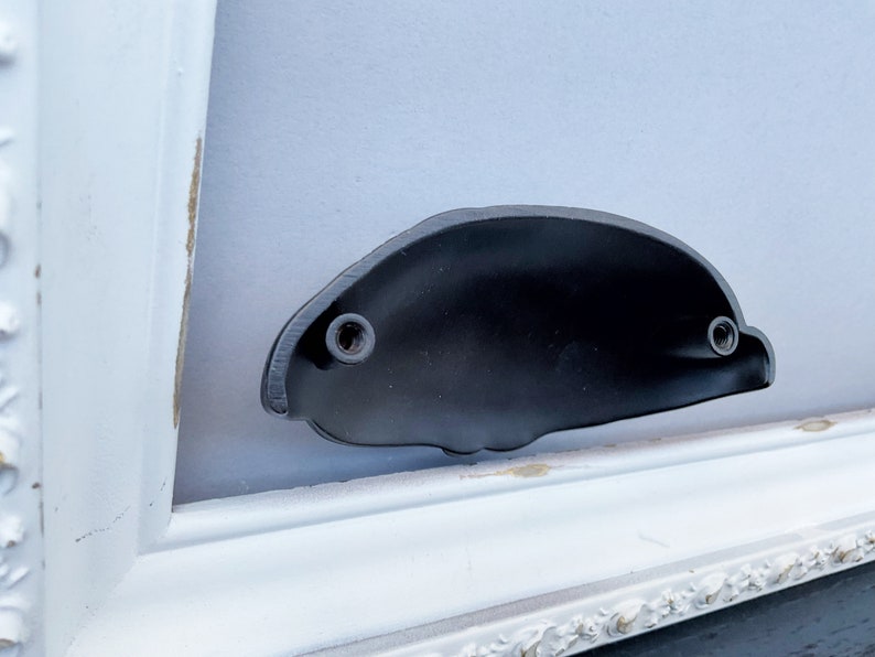 May include: A black metal door handle with two visible screws. The handle is mounted on a white painted frame.