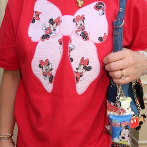 May include: A bright red t-shirt featuring a large, textured, light pink bow appliqu&eacute; patterned with small illustrations of Minnie Mouse. A blue denim bag strap hangs over the shoulder, adorned with a clear charm labeled Cherry Sour Ball.