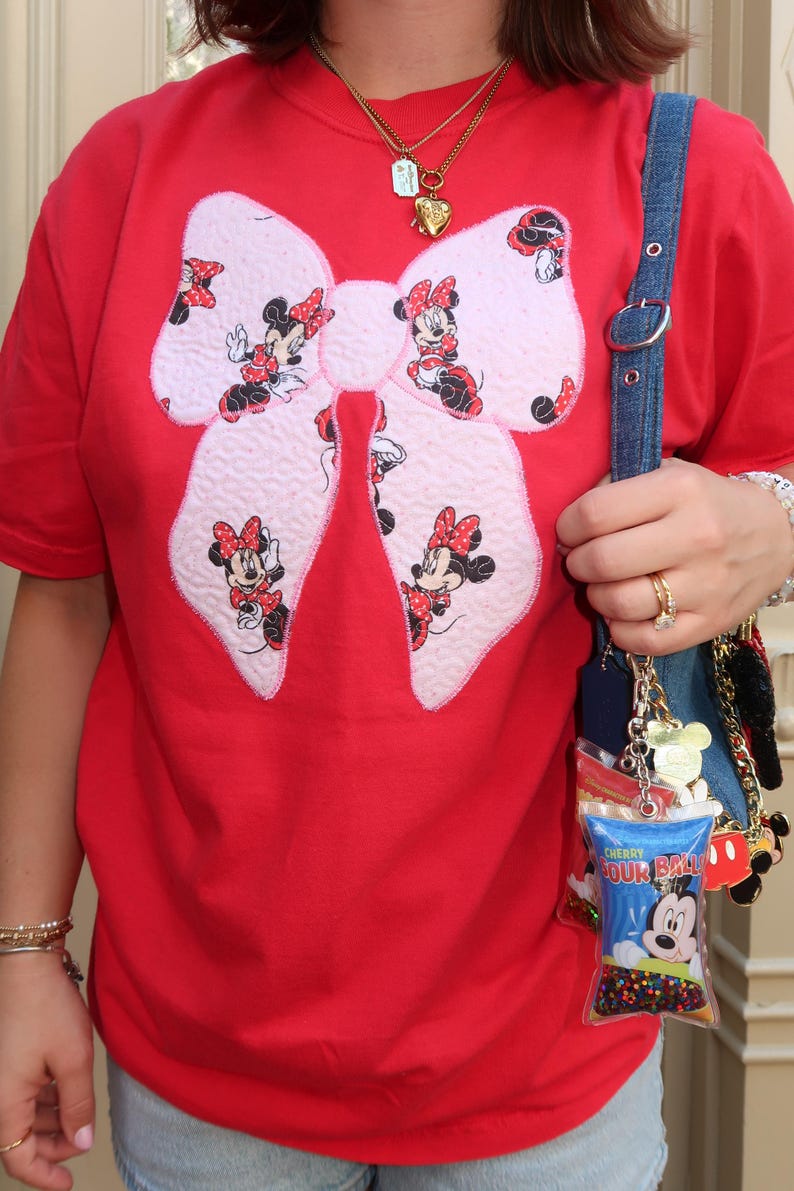 May include: A bright red t-shirt featuring a large, textured, light pink bow appliqu&eacute; patterned with small illustrations of Minnie Mouse. A blue denim bag strap hangs over the shoulder, adorned with a clear charm labeled Cherry Sour Ball.