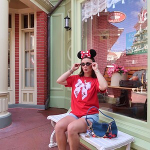 May include: A red t-shirt featuring a large white bow graphic with a Minnie Mouse pattern, light blue denim shorts, white athletic sneakers, black sunglasses, and a blue denim shoulder bag with various small character charms attached to the strap.