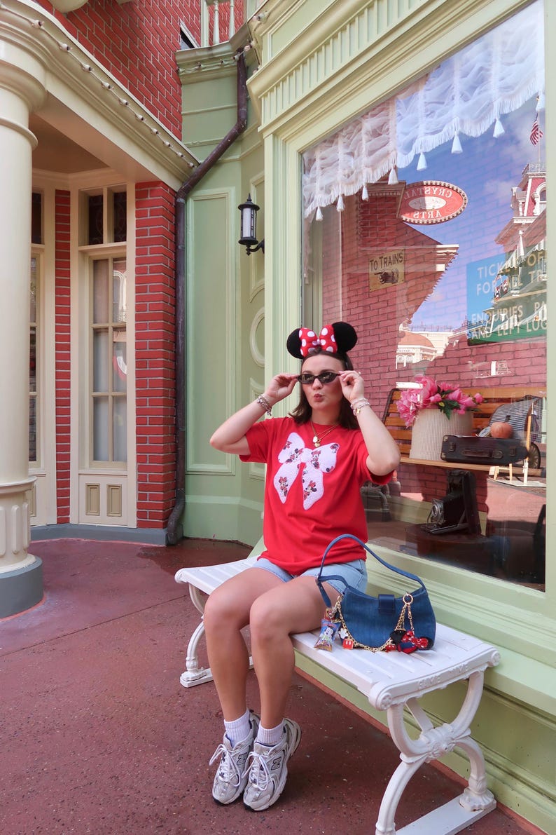 May include: A red t-shirt featuring a large white bow graphic with a Minnie Mouse pattern, light blue denim shorts, white athletic sneakers, black sunglasses, and a blue denim shoulder bag with various small character charms attached to the strap.