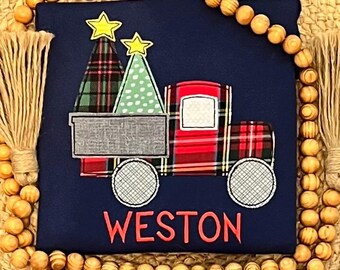 May include: Navy blue Christmas-themed applique with a red plaid truck carrying two Christmas trees. One tree is green with white polka dots, and the other is plaid. The name "WESTON" is embroidered in red below the truck.