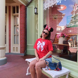 May include: A red short-sleeved t-shirt featuring a large white bow graphic with a floral pattern, paired with blue denim shorts, white socks, grey sneakers, a dark blue denim shoulder bag with charms, and black Minnie Mouse ears with a red polka dot bow.