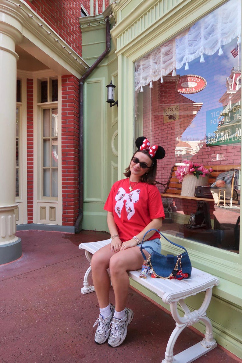 May include: A red short-sleeved t-shirt featuring a large white bow graphic with a floral pattern, paired with blue denim shorts, white socks, grey sneakers, a dark blue denim shoulder bag with charms, and black Minnie Mouse ears with a red polka dot bow.