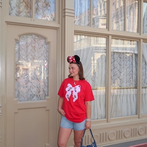 May include: A red t-shirt featuring a large white bow graphic with a Minnie Mouse pattern, light blue denim shorts, white socks, silver sneakers, a blue denim shoulder bag with various character charms, and black Minnie Mouse ears with a red polka dot bow.