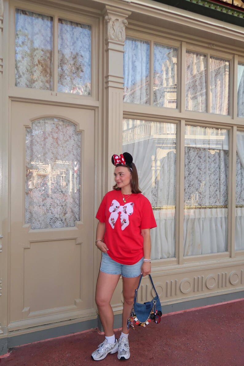 May include: A red t-shirt featuring a large white bow graphic with a Minnie Mouse pattern, light blue denim shorts, white socks, silver sneakers, a blue denim shoulder bag with various character charms, and black Minnie Mouse ears with a red polka dot bow.