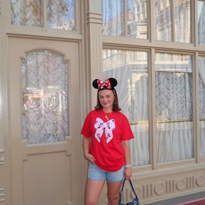 May include: A bright red short-sleeved t-shirt featuring a large white bow graphic with a Minnie Mouse pattern, paired with light blue denim shorts, white sneakers, and a blue denim shoulder bag adorned with various colorful character charms.
