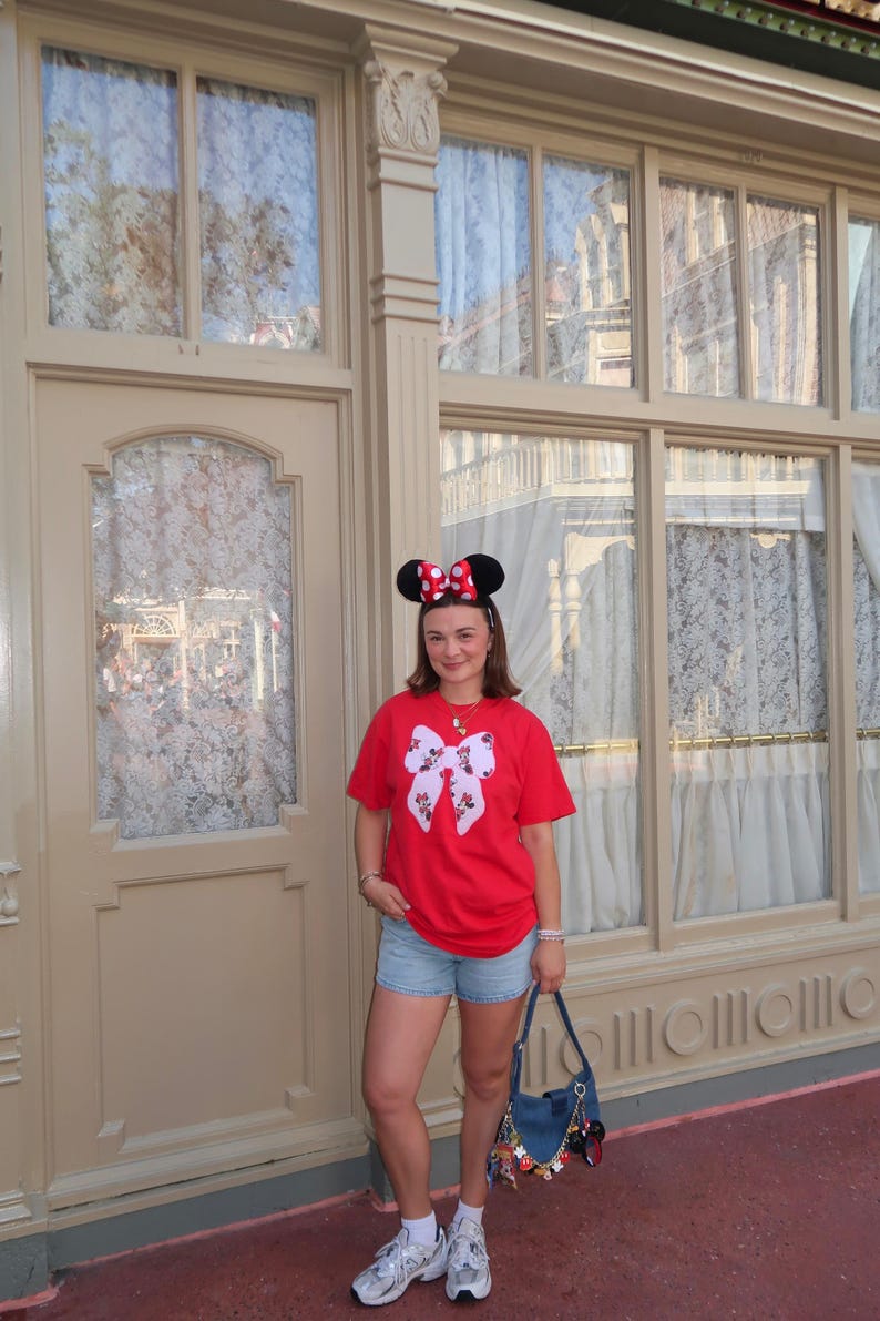 May include: A bright red short-sleeved t-shirt featuring a large white bow graphic with a Minnie Mouse pattern, paired with light blue denim shorts, white sneakers, and a blue denim shoulder bag adorned with various colorful character charms.