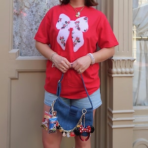 May include: A bright red short-sleeved t-shirt featuring a large white bow graphic with Minnie Mouse patterns. Paired with light blue denim shorts and a dark blue denim shoulder bag adorned with a gold chain and various character-themed charms.