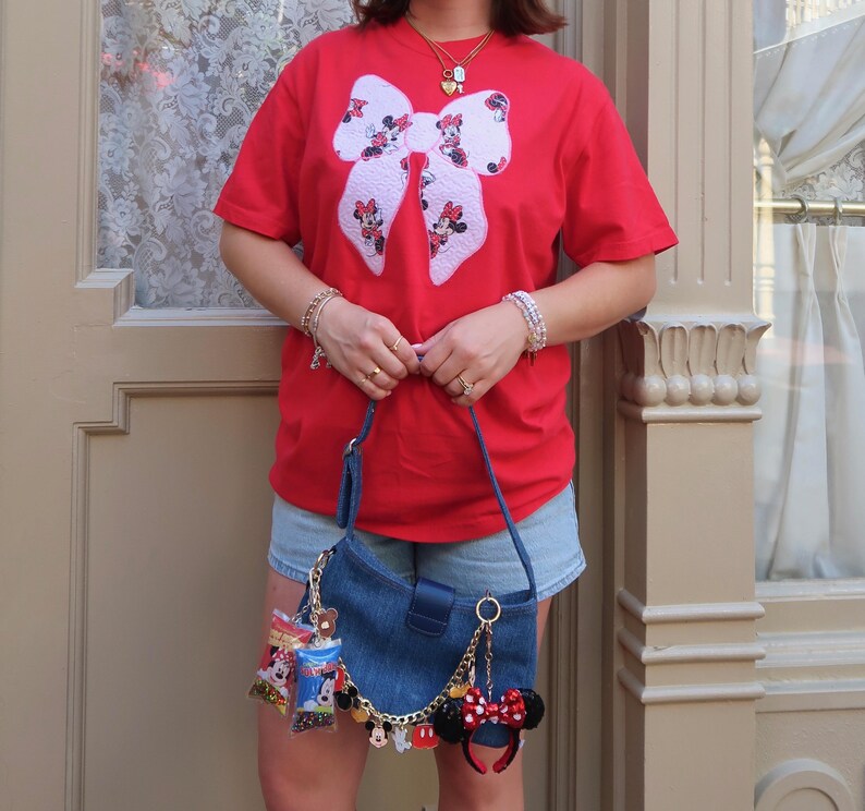 May include: A bright red short-sleeved t-shirt featuring a large white bow graphic with Minnie Mouse patterns. Paired with light blue denim shorts and a dark blue denim shoulder bag adorned with a gold chain and various character-themed charms.
