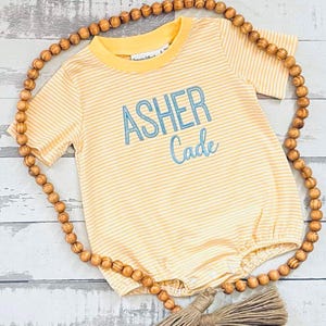 May include: A yellow and white striped baby onesie with the name "Asher Cade" embroidered in blue thread.