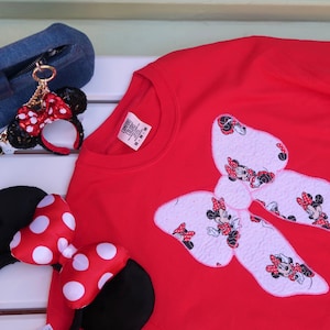 May include: A bright red crewneck t-shirt featuring a large white bow applique patterned with Minnie Mouse illustrations. The shirt is accompanied by a pair of black headband ears with a red and white polka dot bow and a small matching keychain.