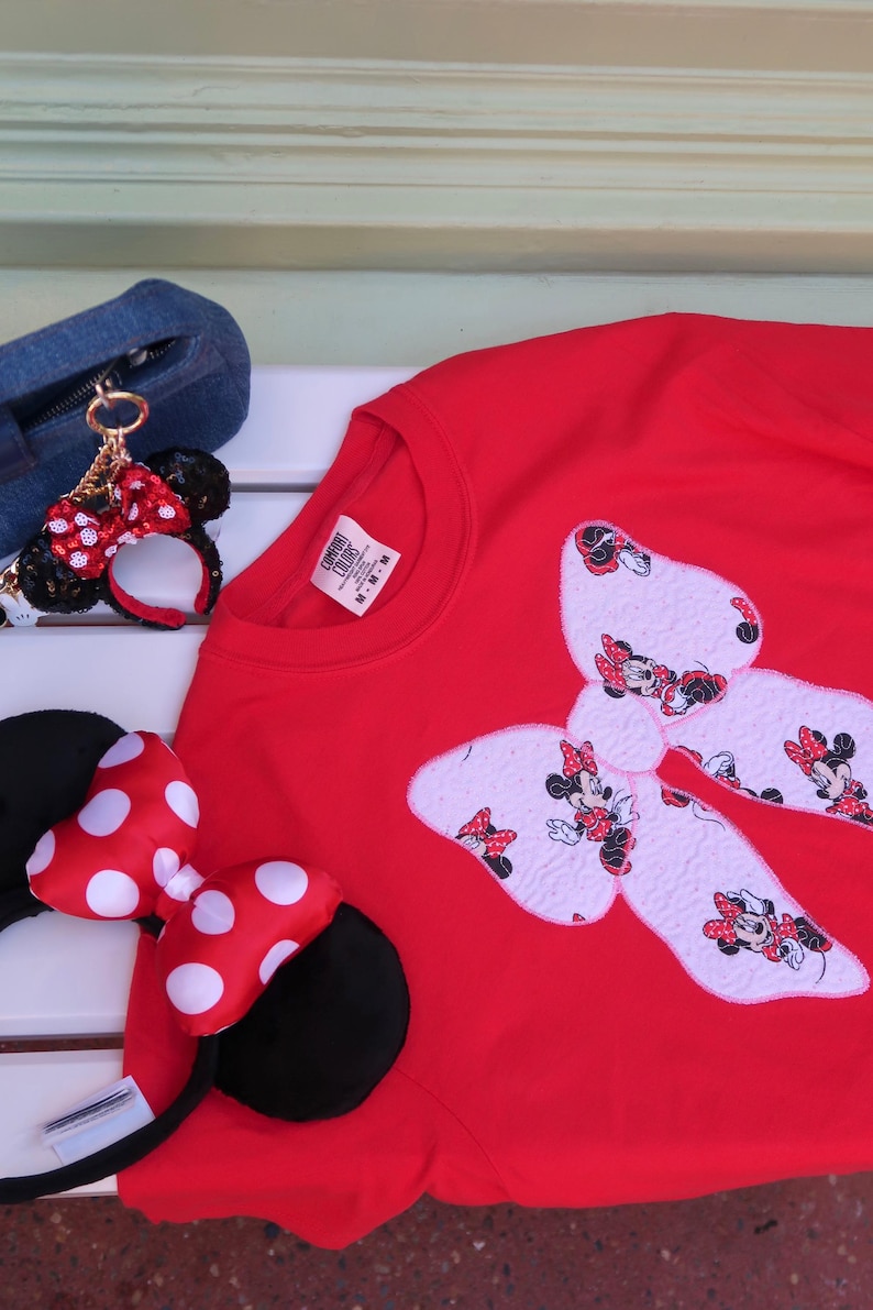 May include: A bright red crewneck t-shirt featuring a large white bow applique patterned with Minnie Mouse illustrations. The shirt is accompanied by a pair of black headband ears with a red and white polka dot bow and a small matching keychain.