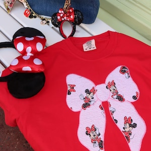 May include: A red crewneck t-shirt featuring a large light pink bow applique with a repeating Minnie Mouse print. The shirt is accompanied by a blue denim handbag with a gold chain charm and a pair of black headband ears with a red polka dot bow.