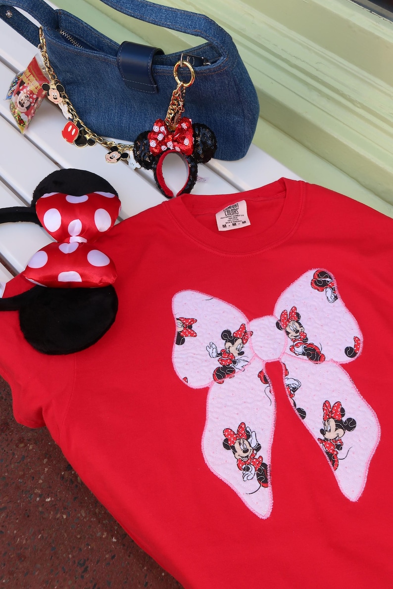 May include: A red crewneck t-shirt featuring a large light pink bow applique with a repeating Minnie Mouse print. The shirt is accompanied by a blue denim handbag with a gold chain charm and a pair of black headband ears with a red polka dot bow.