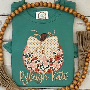 May include: A teal short-sleeved shirt featuring a central pumpkin graphic with a floral pattern in shades of orange, pink, and green, topped with a large cream-colored polka dot bow. The name Ryleigh Kate is embroidered in gold cursive text.
