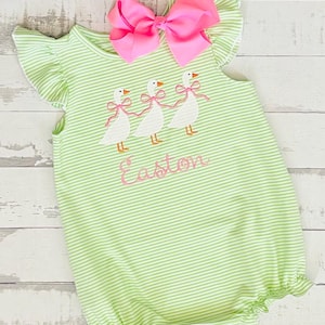May include: A green and white striped romper with ruffle sleeves and a large pink bow. The romper features an embroidered design of three white geese with pink bows and the name "Easton" in pink script.