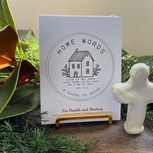 May include: A white card with the words "HOME WORDS" and a house illustration, held by a gold stand, next to a white ceramic cross. The card includes the text "A GUIDE TO ABIDE" and "For Health and Healing".