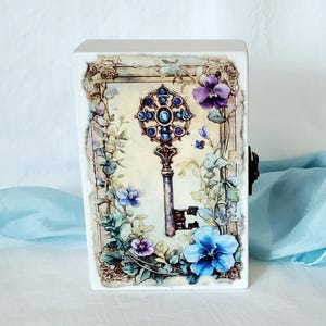 May include: A white rectangular box with a decorative key and floral design. The key is ornate with blue and purple accents, surrounded by purple and blue flowers and green leaves. A small metal clasp is on the side.