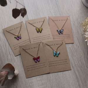 Può includere: Cinque collane d'oro con ciondoli a forma di farfalla su carte marroni che recitano "Make a Wish". Le farfalle sono di diversi colori: rosa, giallo, blu, rosso e turchese.