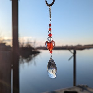 May include: A crystal suncatcher with a red heart and clear beads hanging from a metal hook. The suncatcher is hanging over a body of water.